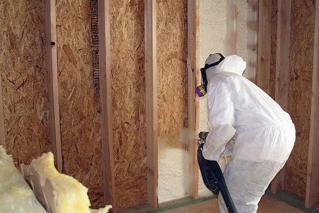 A man in a protective suit is spraying insulation on a wall.