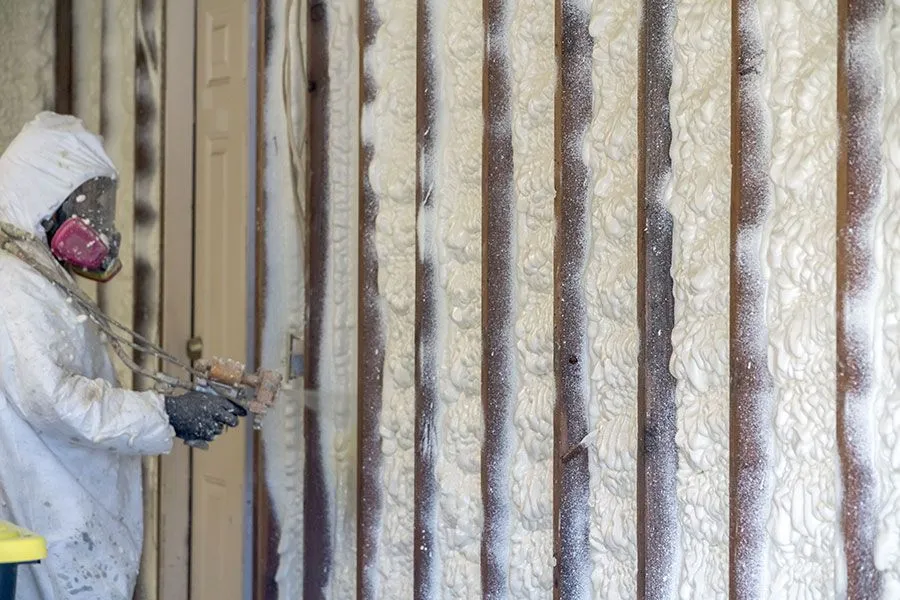 Person in protective suit spraying foam insulation onto wooden wall.