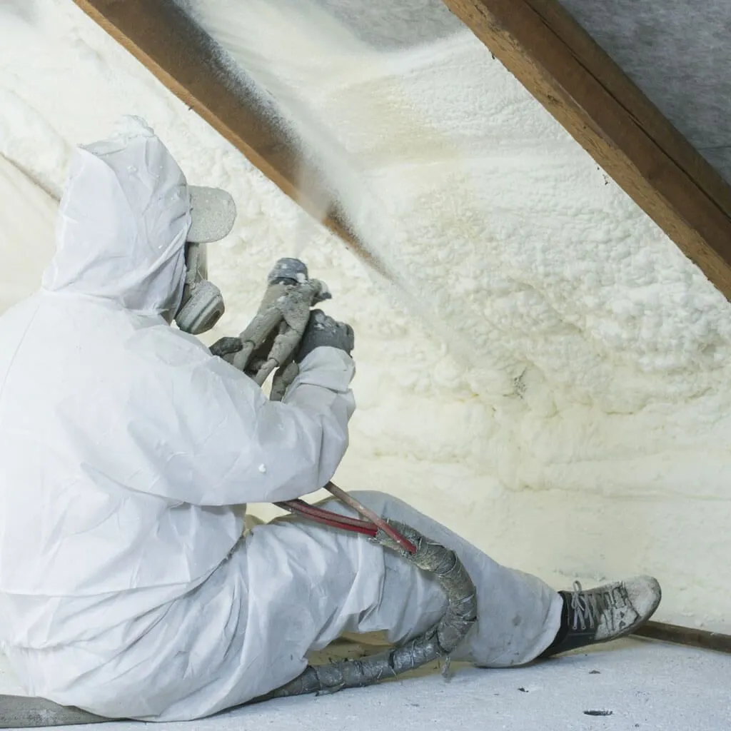 A man in a white suit is spraying insulation in an attic.